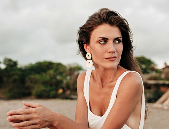 Frau mit langen braunen Haaren und weißem Top sitzt im Freien.